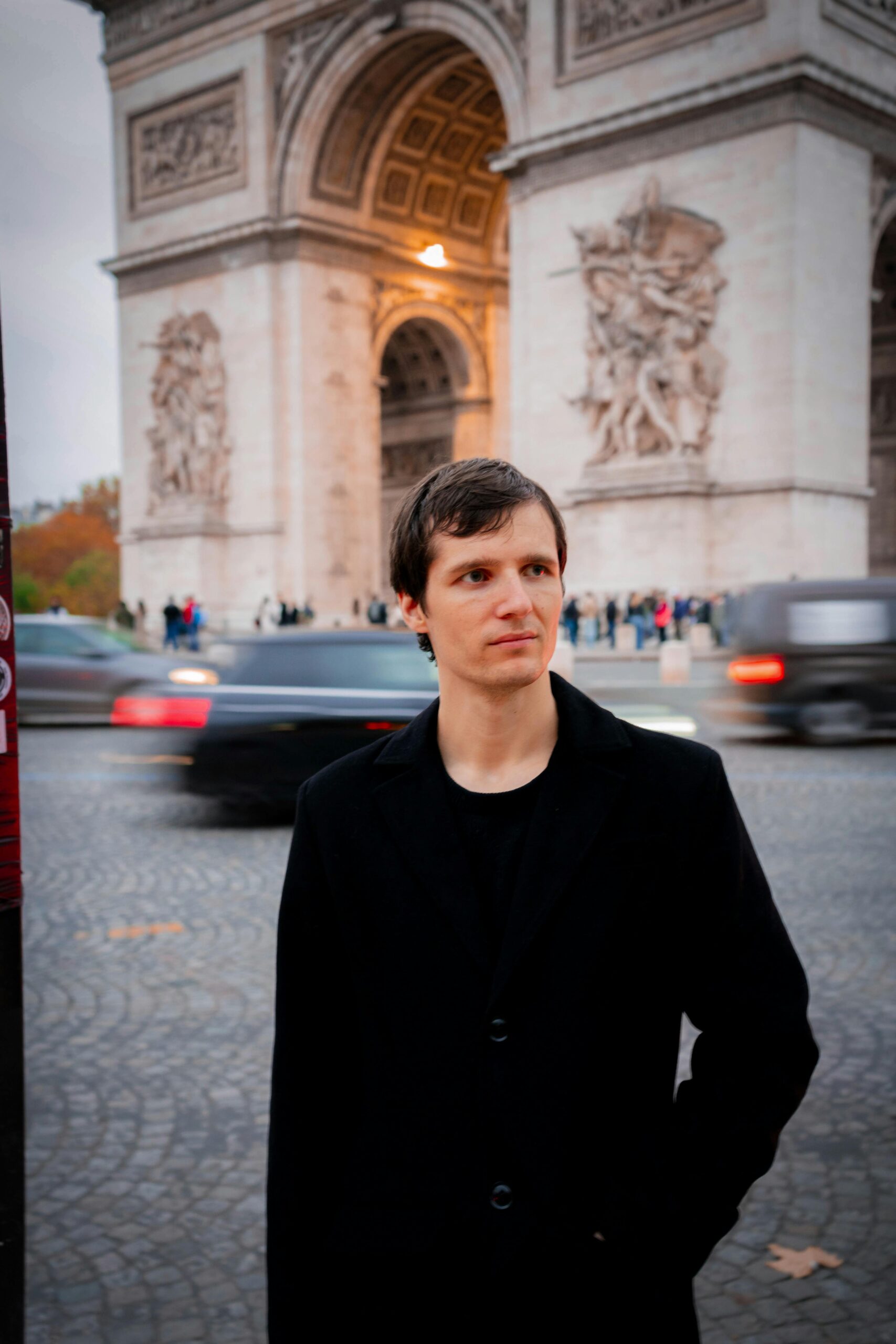 Man wearing a black coat in front of the iconic Arc de Triomphe in Paris during fall.